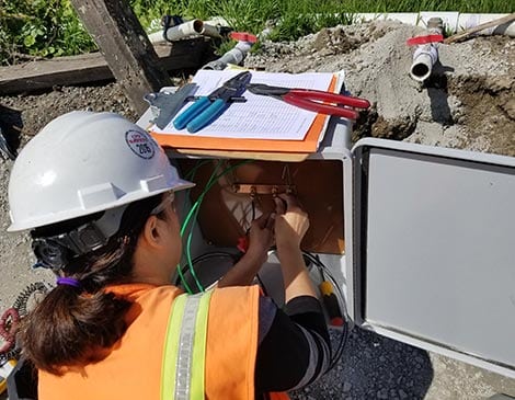 A woman in an orange vest and hard hat working on wires.